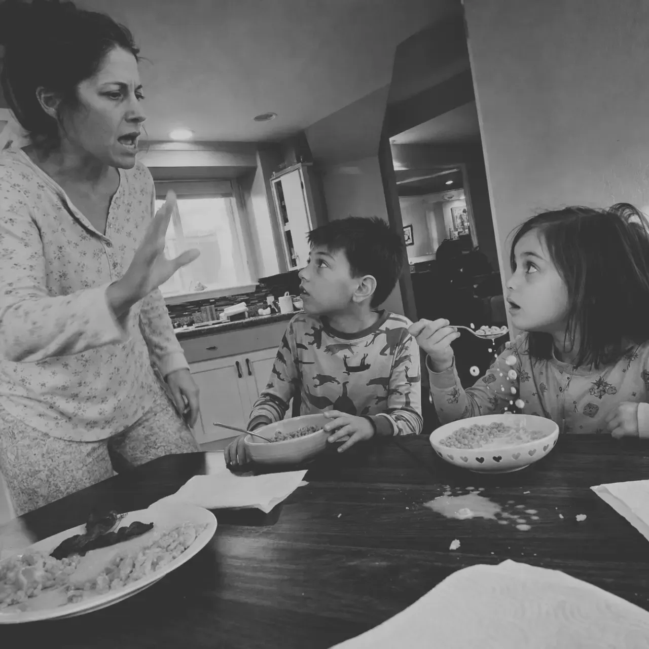 A mother in pajamas stands in the kitchen at seven in the morning, raising her voice at her two children as they eat cereal and look up at her.